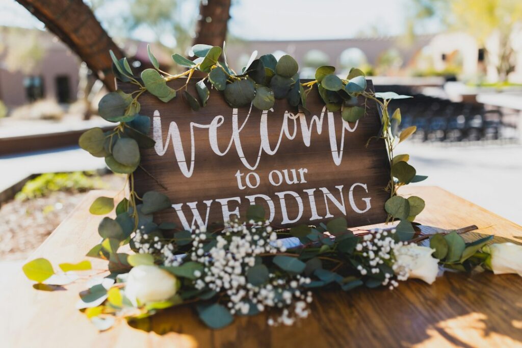 Drewniana tablica Welcome to our Wedding ozdobiona eukaliptusem i białymi kwiatami przy wejściu na wesele